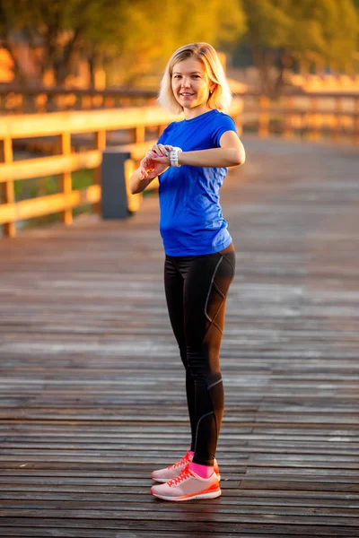 Sportliche Frau blickt auf Uhr, die im Sommer auf Holzbrücke im Park steht . — Stockfoto