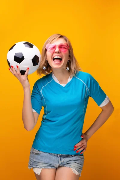 Foto de rubia en gafas de color rosa con pelota de fútbol sobre fondo naranja — Foto de Stock