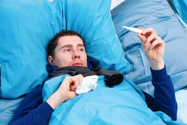 Φωτογραφία του άρρωστου άντρα με κασκόλ με θερμόμετρο στο χέρι που κείτεται στο κρεβάτι — Φωτογραφία Αρχείου