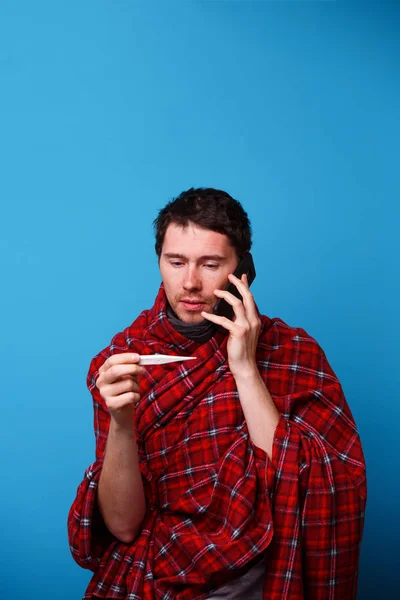 Un hombre enfermo envuelto en una manta está de pie con un termómetro en sus manos y llamando al médico . — Foto de Stock