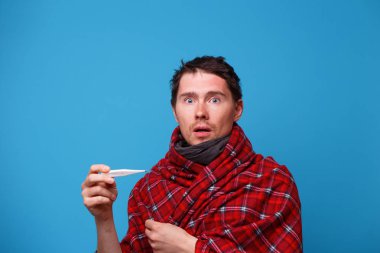 Battaniyeye sarılmış hasta bir adam elinde termometreyle duruyor..