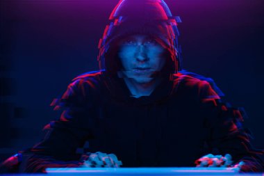 Kapüşonlu hacker karanlık odada bilgisayar yazma metni ile çalışıyor. Glitch efektli görüntü