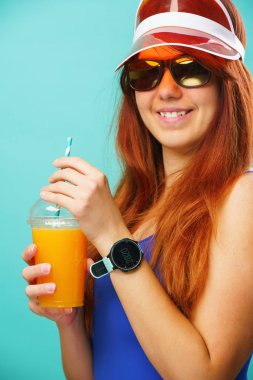 Mavi mayo, şapka ve güneş gözlüğü giyen kadın bir bardaktan meyve suyu içer