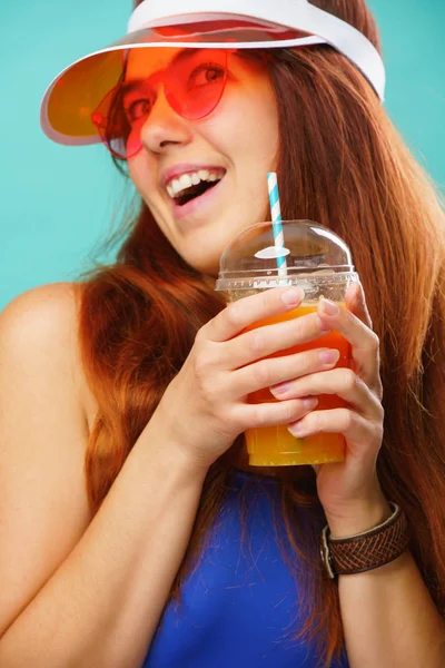 Mavi mayo, şapka ve güneş gözlüğü giyen kadın bir bardaktan meyve suyu içer