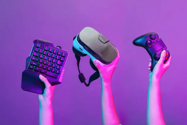 Set of hands with gamepad, keyboard, virtual reality headset and headphones on violet background. — Stock Photo, Image