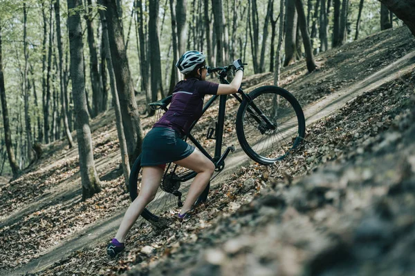 Wizerunek kobiecej sportowca w kasku podnosząc rower na wzgórze w lesie — Zdjęcie stockowe