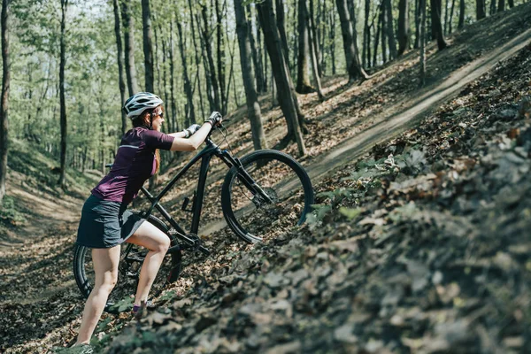 Zdjęcie kobiecej sportowca w kasku podnosząc rower na wzgórze w lesie — Zdjęcie stockowe