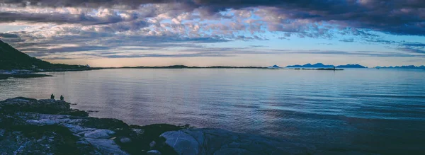 Foto panorâmica de pessoas sentadas em uma praia rochosa ao pôr do sol na Noruega — Fotografia de Stock