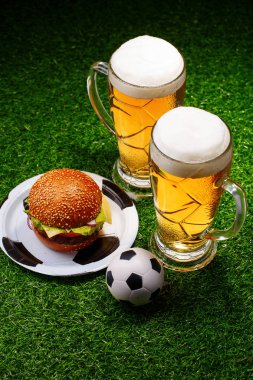 Yeşil çimenler üzerinde iki bardak bira, hamburger ve futbol topu.