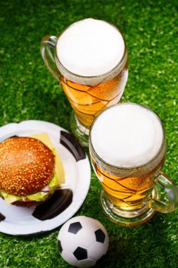 Yeşil çimenler üzerinde iki bardak bira, hamburger ve futbol topu.