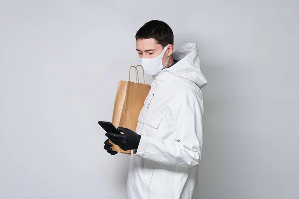 Hombre con una máscara protectora para evitar la propagación de Coronavirus bolsa de la compra y se comunica a través de un teléfono inteligente . — Foto de Stock