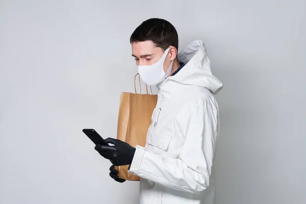 Homem usando uma máscara protetora para evitar a propagação do Coronavirus segurar saco de compras e se comunica via smartphone . — Fotografia de Stock