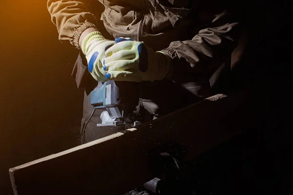 Photo of a worker in outfit with blue gloves working with electrical jigsaw on black background with upper light and front view.