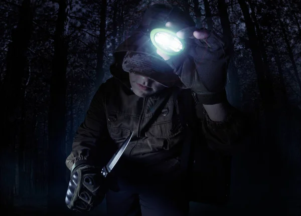 Profile view photo of a male person in brown tactical outfit jacket, gloves and knife turning on head flashlight on night woods background.