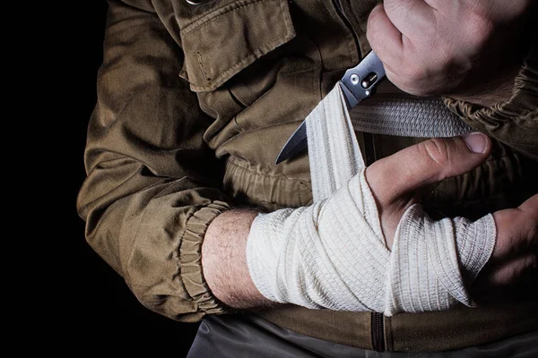 Profile view photo of a male person in brown tactical outfit jacket taking out bandage and cutting it with knife on black background.