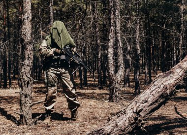 Tam teçhizatlı bir orman keskin nişancısının fotoğrafı. Tüfekli ve taktik atkısı ormanda duruyor..