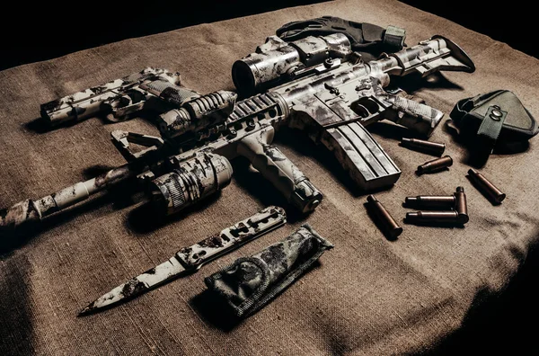 Photo of a camouflaged military AR-15 rifle with scope laying with knife, gun and tactical gloves side view.