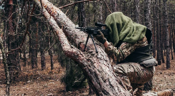 图为一名全副武装 手持来复枪 手持伪装的森林狙击手坐在树林中的一棵树旁瞄准的照片 — 图库照片