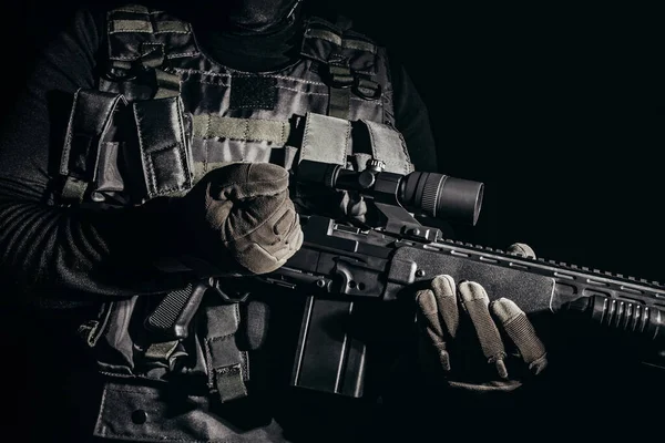 Photo of a soldier in tactical gloves and armor vest standing and reloading sniper rifle on black background.