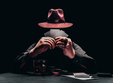 Siyah gömlekli ve şapkalı bir gangsterin fotoğrafı. Poker oynuyor ve elinde oyun fişleri ve parayla siyah kart tutuyor..