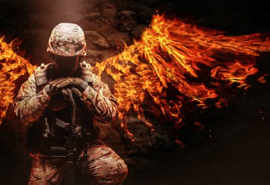 Tam teçhizatlı bir askerin fotoğrafı, miğfer, çelik yelek ve tüfekle arkasında ateş kanatları ile diz çökmüş..