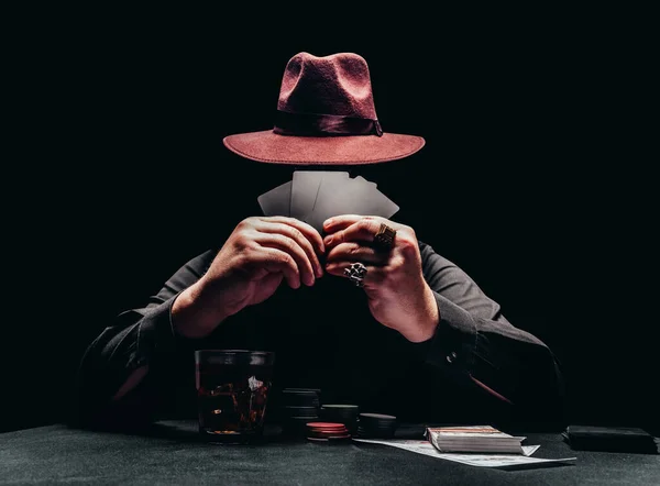 Фотографії затіненого гангстера в чорній сорочці та капелюх, що грає в покер, і тримають чорні картки з чіпсами та грошима. Ліцензовані запасні зображення