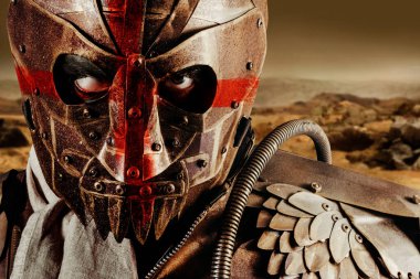 Çölde duran, metal zırh ve çelik maskeli, kırmızı haçlı, kıyamet sonrası yağmacı savaşçısının fotoğrafı..