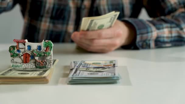 銀行券を数えてる 新しい購入のためのお金を数える 旅のためのお金を節約 貯蓄の概念 何千ドルものテーブルの上に小さな家 シェルがあります — ストック動画