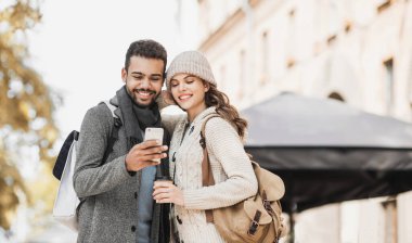Akıllı telefon kullanan mutlu güzel bir çift. Genç, neşeli bir kadın ve bir adam sonbaharda bir şehirde cep telefonuna bakıyor. Teknoloji, seyahat, turizm, öğrenci konsepti