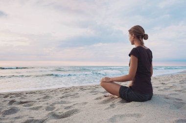 Dışarıda yoga yapan genç bir kadın. Arka planda gün batımı manzaralı meditasyon yapan bir kız. Uyum, meditasyon, rahatlama, tatiller, özgürlük, sağlıklı yaşam tarzı konsepti.