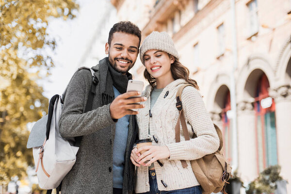 Beautiful happy couple using smartphone. Young joyful smiling woman and man looking at mobile phone in a city in autumn. Technology, travel, tourism, students concept