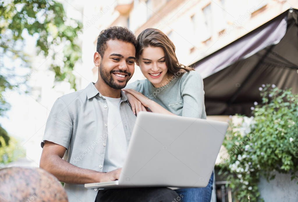 Beautiful happy couple using laptop computer outdoors. Joyful smiling woman and man looking at gadget in city in summer. Love, relationship, technology and communication concept
