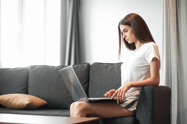 Young woman with modern laptop sitting on sofa at home. Happy girl browsing through the internet during free time..