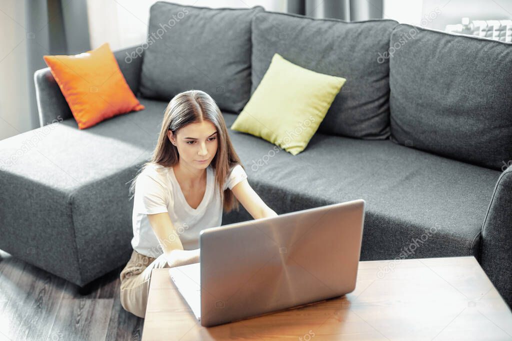 Young woman with modern laptop sitting on sofa at home. Happy girl browsing through the internet during free time.. 