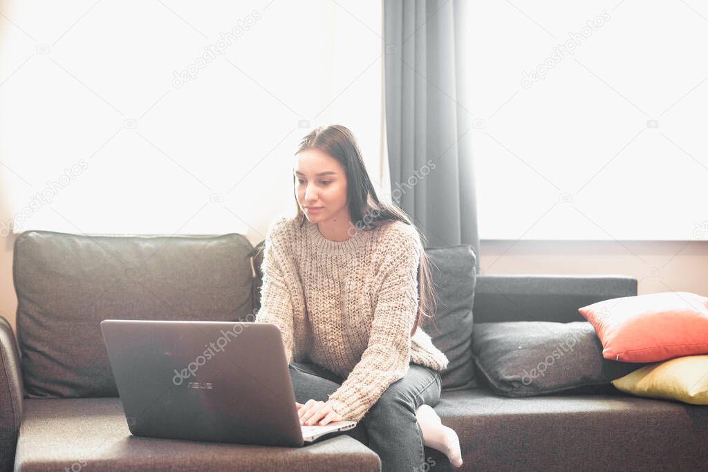 Young woman with modern laptop sitting on sofa at home. Happy girl browsing through the internet during free time.. 