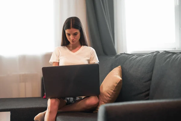 Young woman with modern laptop sitting on sofa at home. Happy girl browsing through the internet during free time..