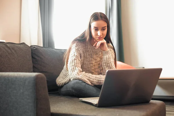 Young woman with modern laptop sitting on sofa at home. Happy girl browsing through the internet during free time..