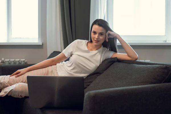 Young woman with modern laptop sitting on sofa at home. Happy girl browsing through the internet during free time..