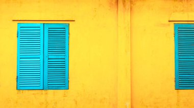 Güneşli şehir ortamında mavi pencereli canlı sarı duvar.