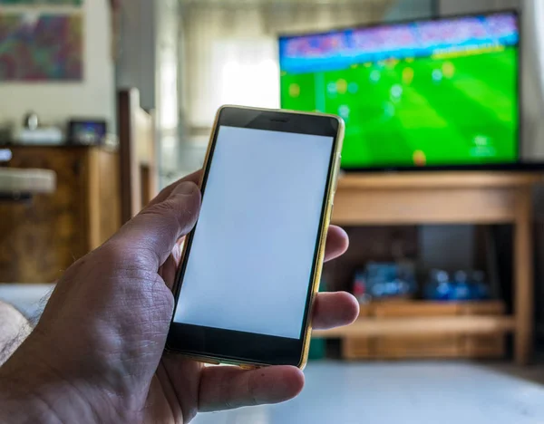 Erkek el closeup smartphone ile beyaz ekran televizyonda futbol maç sırasında kullanır