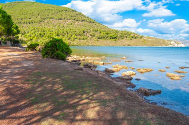 Capocaccia sahilinde, güneşli ve bulutlu bir günde Tramariglio körfezi, iskele ve plaj manzarası