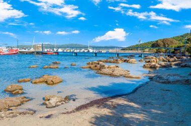 Capocaccia sahilinde, güneşli ve bulutlu bir günde Tramariglio körfezi, iskele ve plaj manzarası