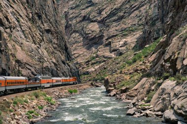 Tren Colorado 'daki Arkansas nehrinin yanındaki kraliyet geçidinin derinliklerine gidiyor. Etrafı kayalık kayalıklarla çevrili mavi suyun yanında turuncu motor.