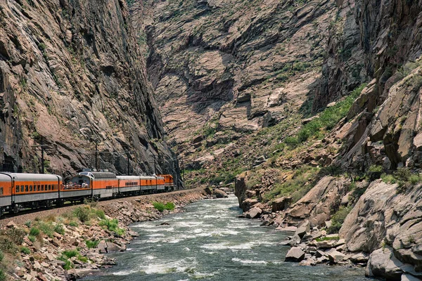 Tren Colorado 'daki Arkansas nehrinin yanındaki kraliyet geçidinin derinliklerine gidiyor. Etrafı kayalık kayalıklarla çevrili mavi suyun yanında turuncu motor.