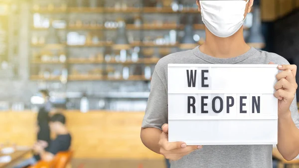 Waiter with protective face mask holding we re open sign while standing at cafe.