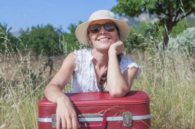 Genç bir kadının yaz mevsiminde elinde eski bir kırmızı bavul tutan şapka ve gözlüklerle tarlada yürümesi, mutluluğu ifade ediyor ve tatilde..