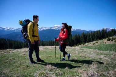 İki yürüyüşçü bir dağ sırtında mola veriyor. Sırt çantalı sportif çift, karlı dağların arka planında duruyorlar..