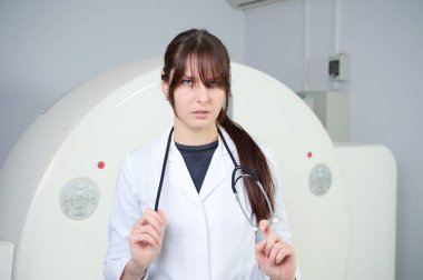 MRI tarayıcısının önünde duran kendinden emin kadın teknisyenin portresi. Beyaz laboratuvar önlüklü kadın tomografinin yanında duruyor. Tarama odasında güzel bir doktor var. Üniformalı Radyolog.