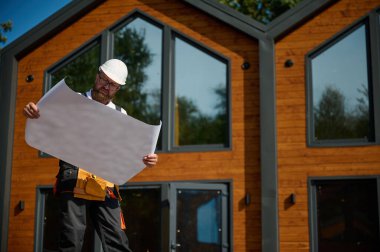 Mimar bekleme planı. Sakallı mimar elinde proje planları, yeni modern evlerin yanında duruyor ve inşaat alanında bir şeyler arıyor..