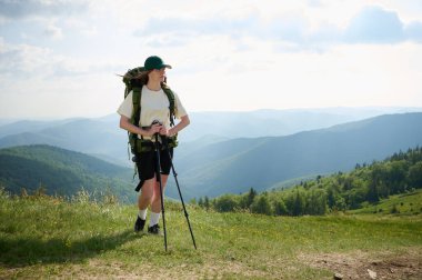 Kadın yürüyüşçü çimenli bir tepede duruyor, engin dağ manzarasını seyrediyor. Şapka ve şort giyen maceraperest, büyük bir sırt çantası taşıyor ve yürüyüş direkleri kullanıyor..